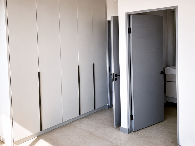 A bright, modern bedroom featuring neutral tiled flooring, extensive built-in cabinetry, and clean finishes, with an adjoining bathroom visible through the doorway.