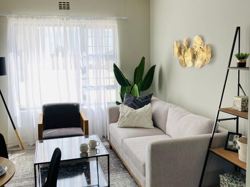 A well-lit living room featuring neutral wall tones, a large window with sheer curtains, and a functional layout suitable for comfortable seating.