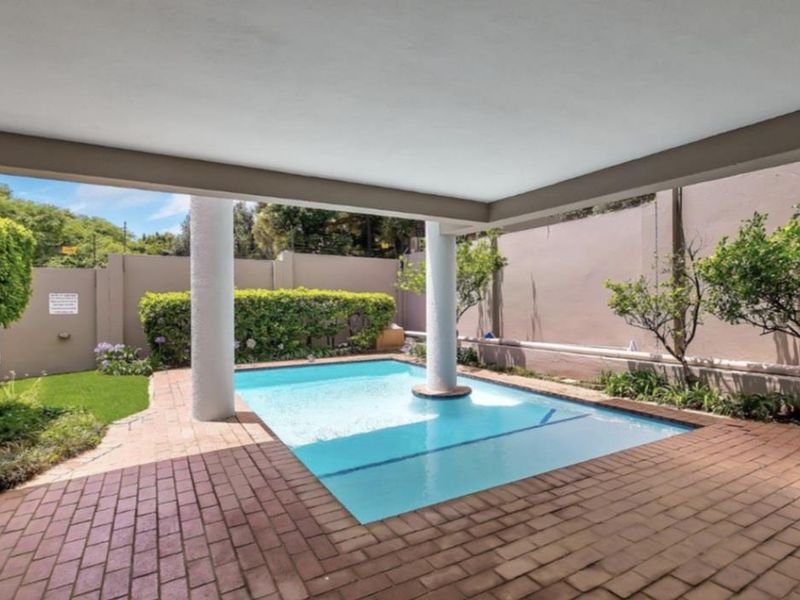 A private swimming pool area featuring a covered patio with brick paving, structural columns, and a surrounding perimeter wall with landscaped garden borders.