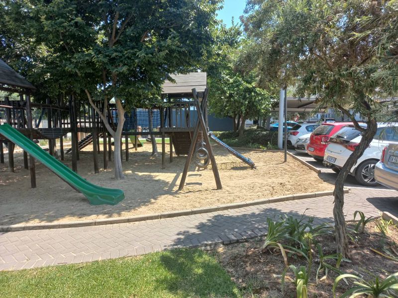 Communal garden area featuring a children's playground with wooden structures and a paved walkway, adjacent to the apartment complex parking area.