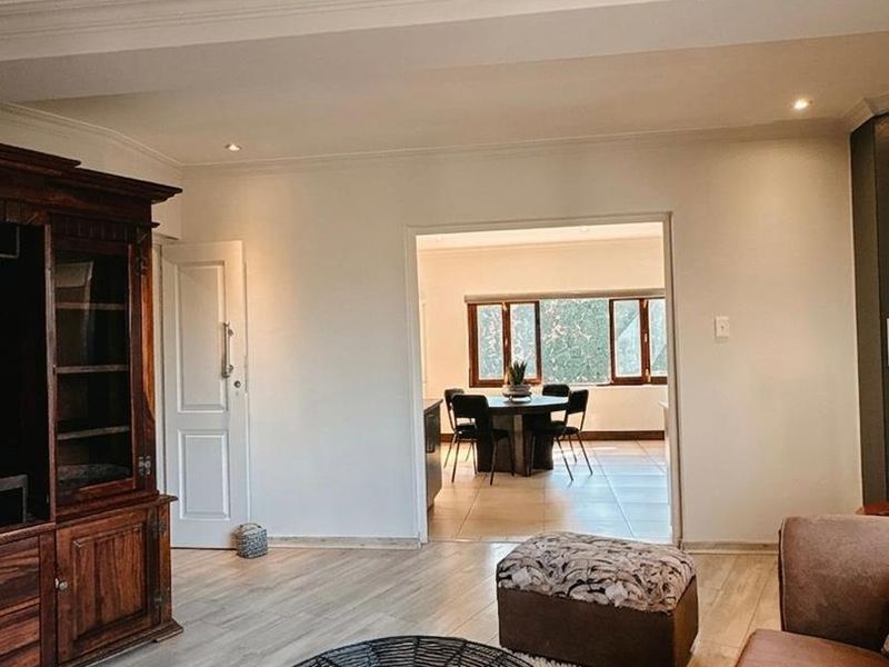 A well-maintained living room featuring laminate flooring, recessed downlighting, and decorative cove cornicing, with an open transition into the adjacent dining area.