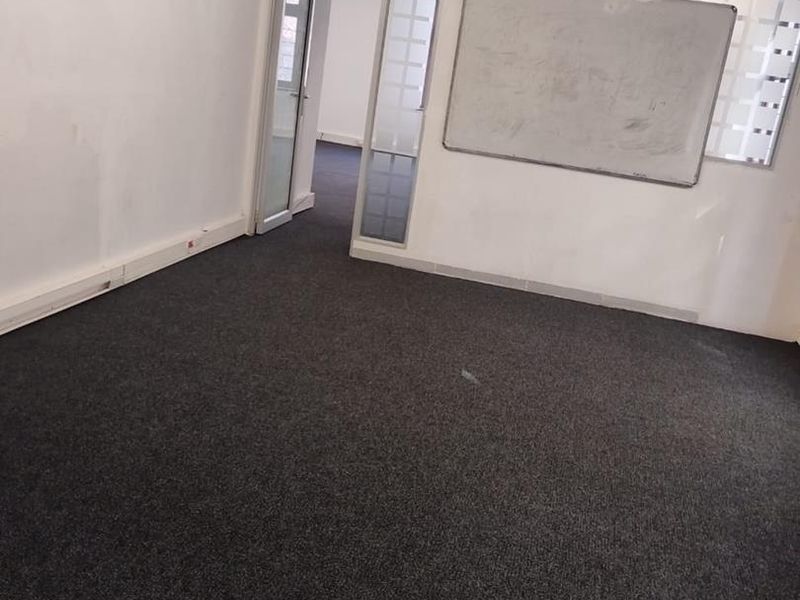 An empty office space featuring dark carpeting, exposed ducting, a glass partition, and a wall-mounted whiteboard.