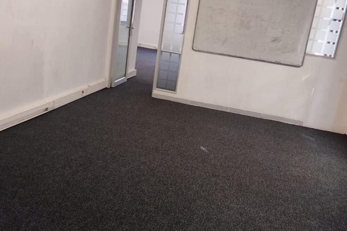 An empty office space featuring dark carpeting, exposed ducting, a glass partition, and a wall-mounted whiteboard.