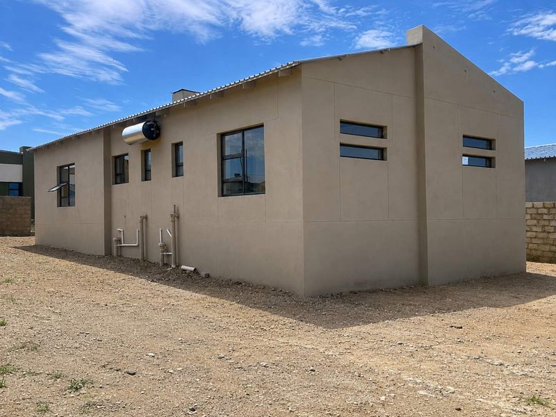 Rear view of a modern house featuring a solar water heater, aluminium window frames, and a metal roof, set on an unfinished plot.