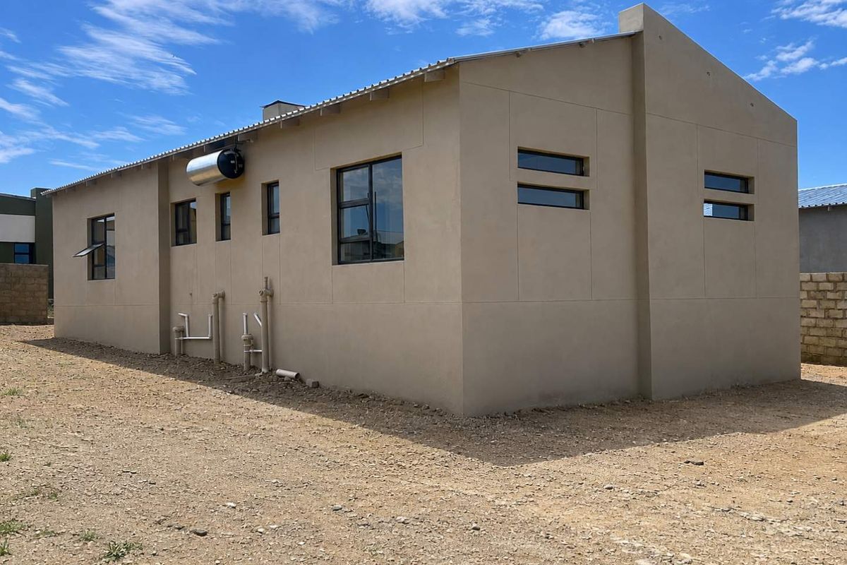 Rear view of a modern house featuring a solar water heater, aluminium window frames, and a metal roof, set on an unfinished plot.