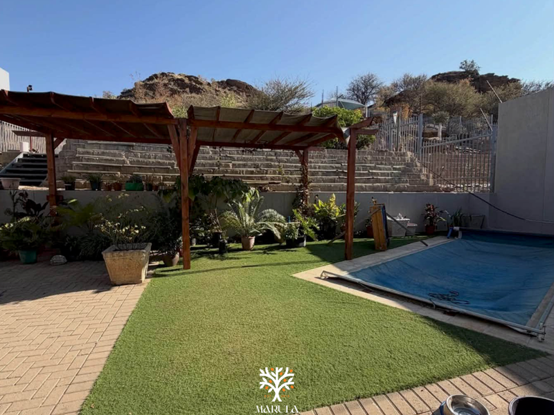 Outdoor pool area featuring a timber pergola, paved patio, artificial lawn, and a retaining wall backdrop, providing a functional space for relaxation.