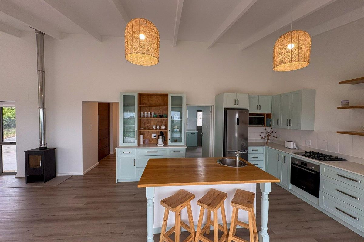 Modern kitchen featuring light green cabinetry, a central wooden island, integrated appliances, and a wood-burning stove under high exposed beam ceilings.
