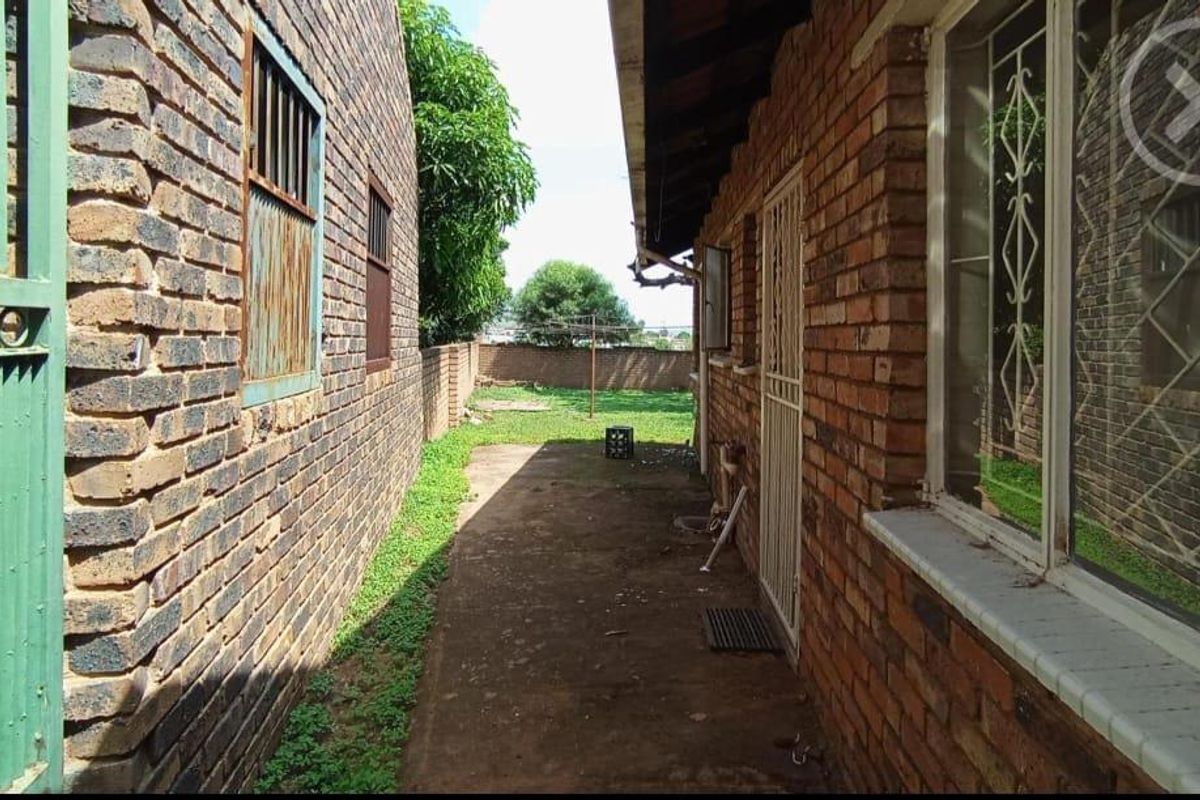 A narrow side exterior pathway featuring exposed brick walls, metal security bars on windows, and a concrete walkway leading to the rear garden area.