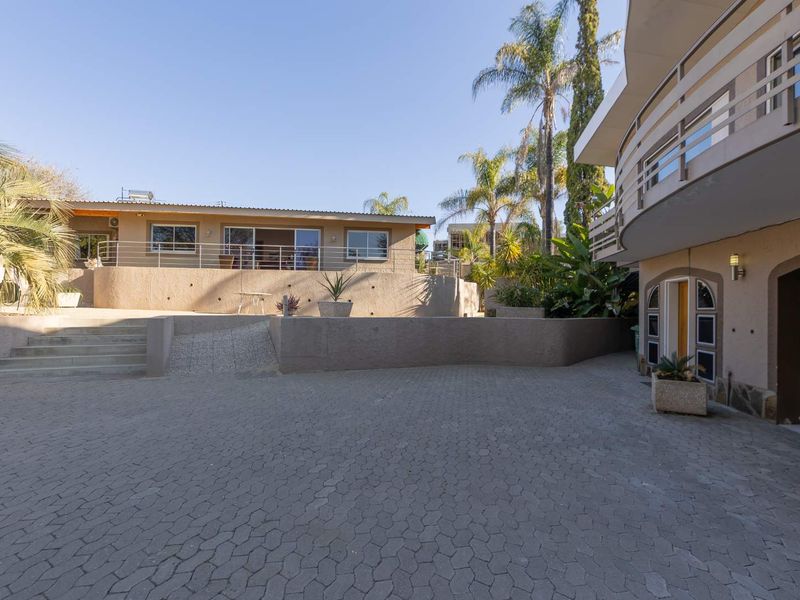 A spacious paved driveway leading to a multi-level residential property featuring a garage, balcony, and landscaped garden areas.
