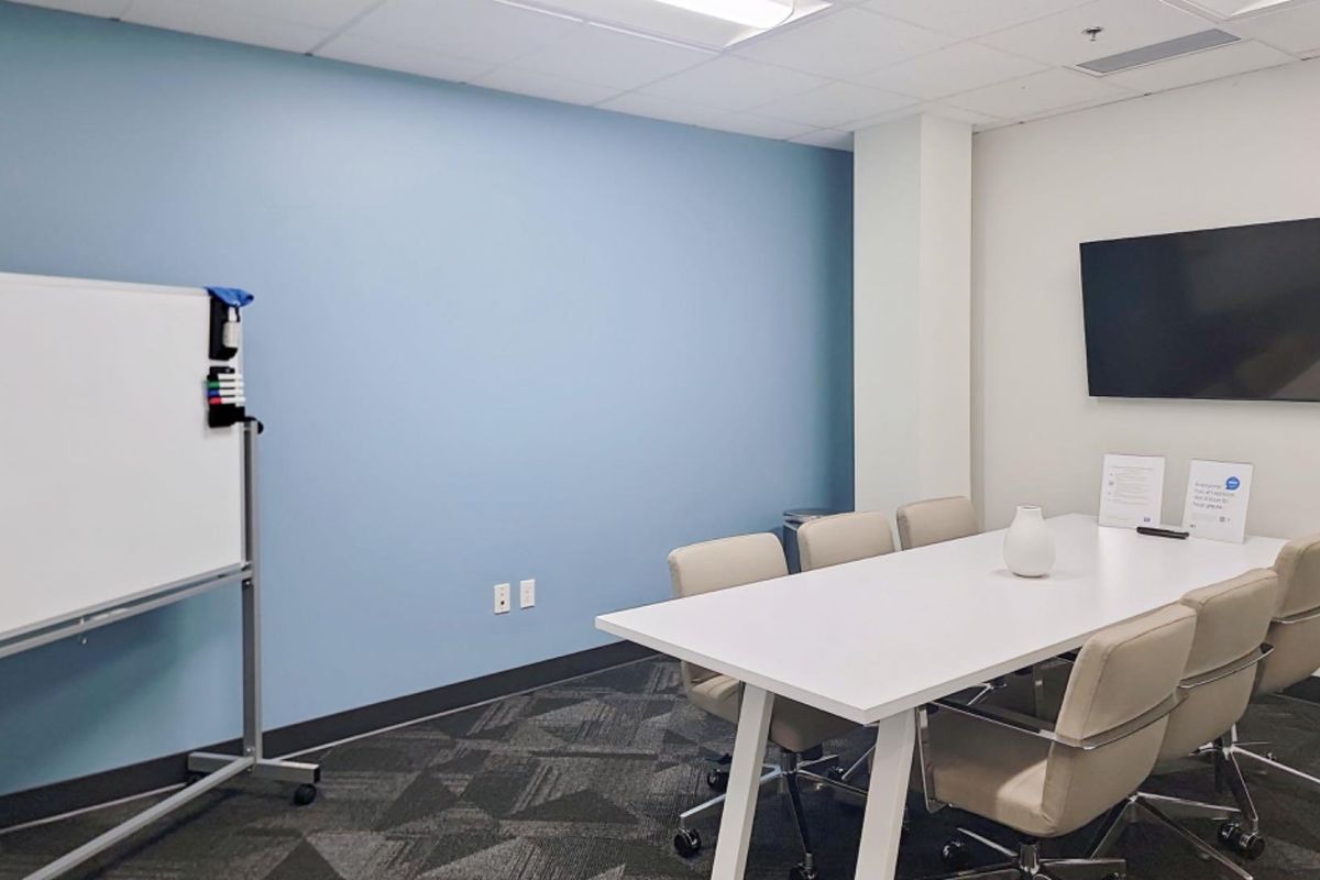 A professional office meeting room featuring a wall-mounted television, carpeted flooring, and a suspended ceiling with recessed lighting.