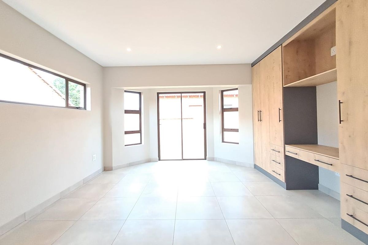 A bright, modern bedroom featuring neutral floor tiles, built-in wooden cabinetry with an integrated desk, and large windows providing ample natural light.