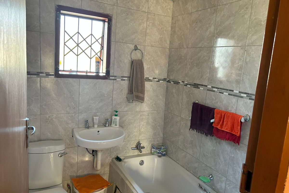 A functional bathroom featuring full-height wall tiling, a standard bathtub, a wall-mounted basin, and a window with security bars.