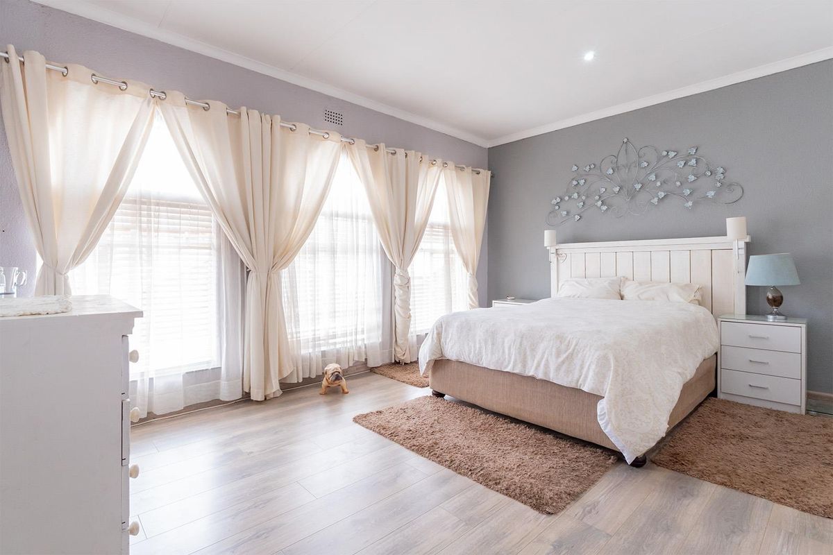 A spacious, well-lit bedroom featuring light-toned laminate flooring, neutral grey walls, and large windows with elegant drapery.