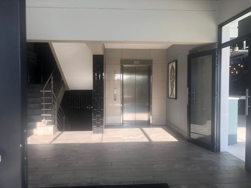 Modern office building entrance lobby featuring a stainless steel elevator, tiled flooring, and a staircase with a contemporary metal handrail.
