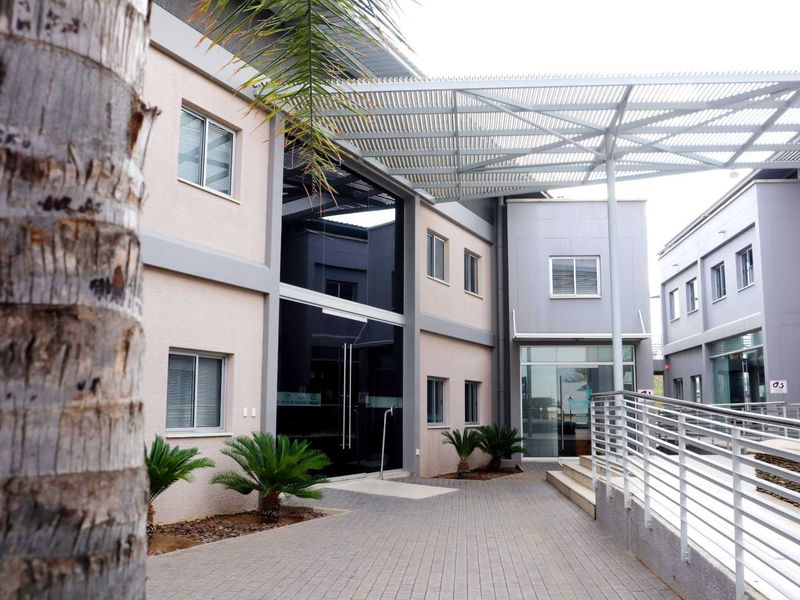 Modern office building exterior featuring a paved entrance walkway, metal pergola structure, and glass facade elements with accessible ramp and stair access.