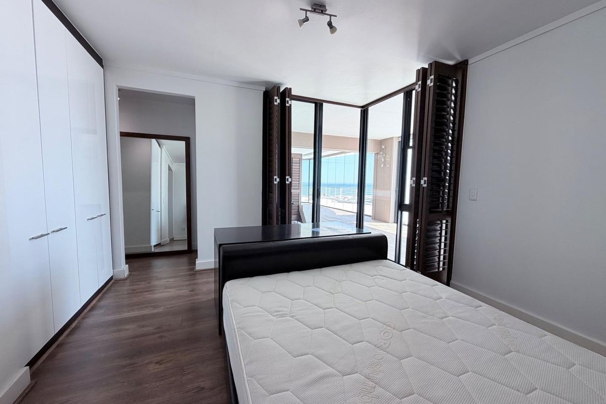 A bright bedroom featuring built-in wardrobes, dark wood-look flooring, and folding shutters opening onto a balcony with ocean views.