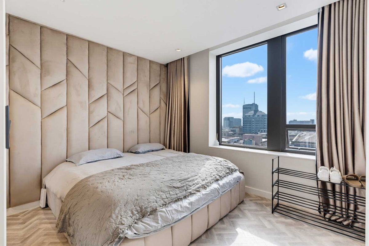 Modern bedroom featuring a custom upholstered feature wall, herringbone flooring, and large windows offering expansive city views.
