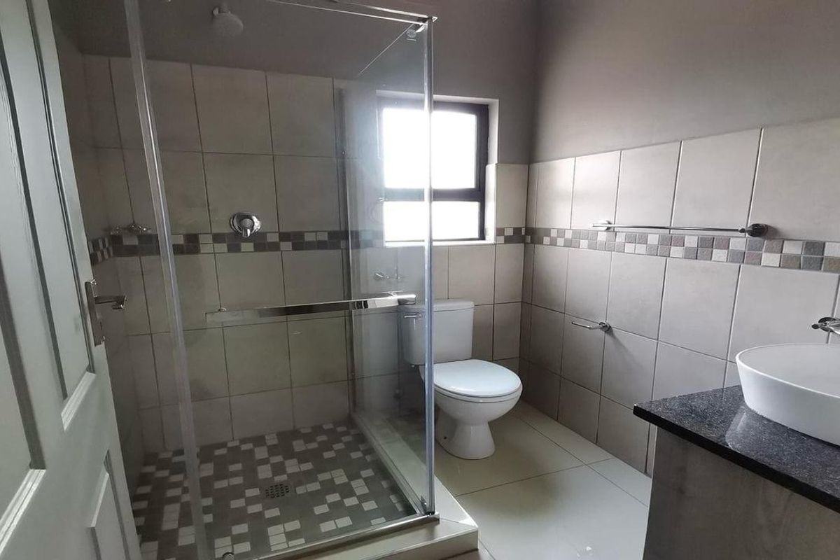 A clean, modern bathroom featuring a glass-enclosed shower, a white toilet, and a vanity basin set into a granite countertop with neutral wall tiling.
