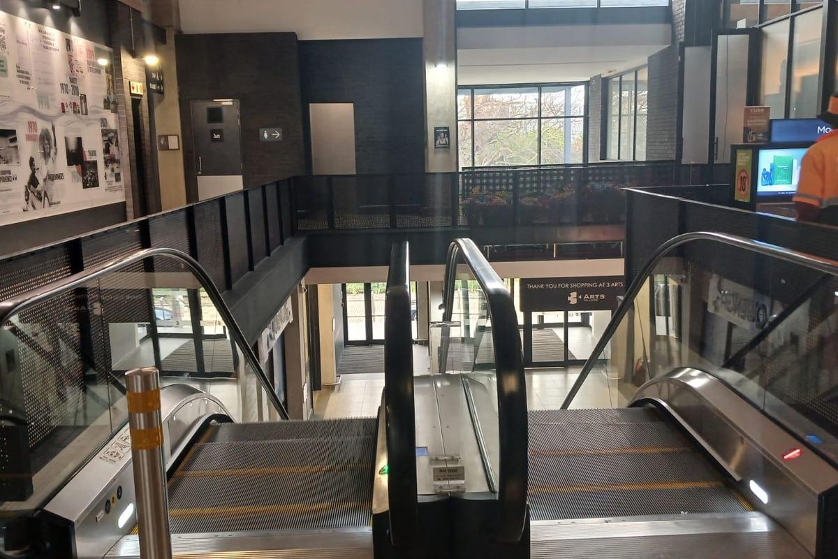 A modern retail center entrance featuring a central escalator, metal balustrades, and clear glass partitions with high ceilings and commercial signage.