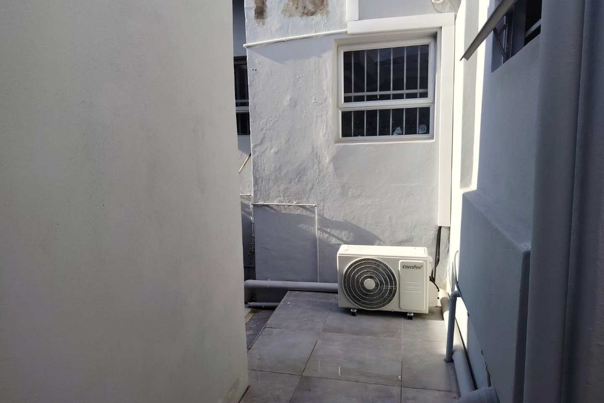 A narrow exterior utility passage featuring tiled flooring, a wall-mounted air conditioning unit, and visible exterior wall maintenance requirements.