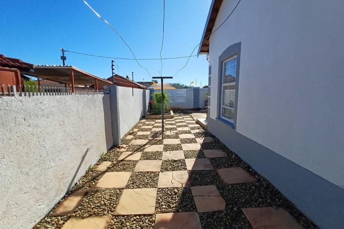 A side exterior area featuring a checkered pattern of paving stones and gravel, enclosed by a boundary wall with electric fencing.