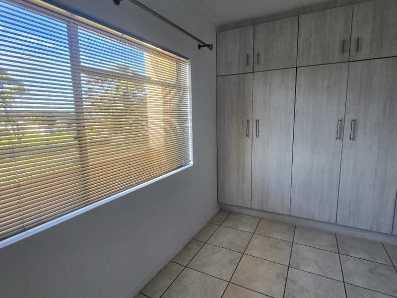 A clean, empty bedroom featuring neutral tiled flooring, ample built-in cupboard storage, and venetian blinds on the window.