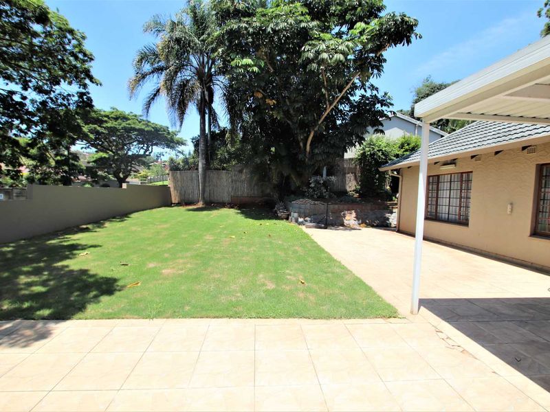 A spacious rear garden featuring a tiled patio, covered veranda, and a well-maintained lawn enclosed by a boundary wall.