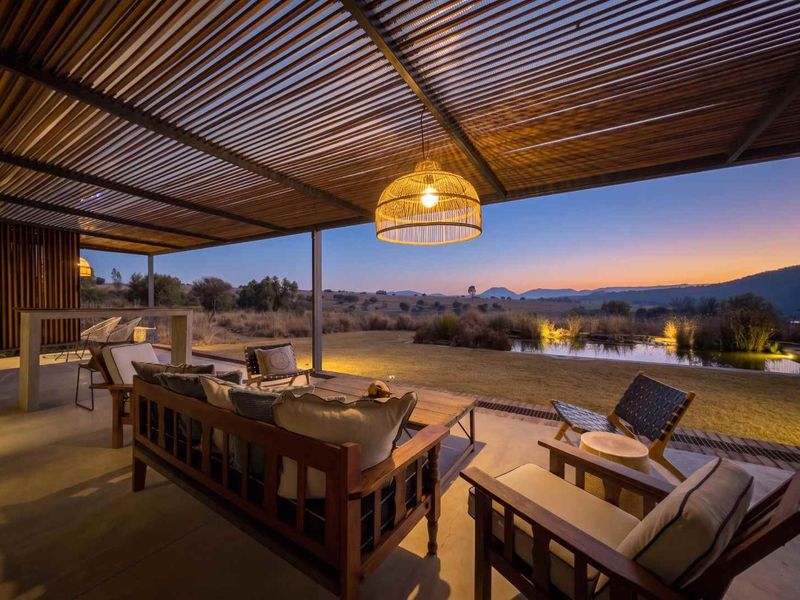 A spacious covered patio featuring a slatted timber pergola, concrete flooring, and a built-in bar, overlooking a scenic landscape and pond at sunset.