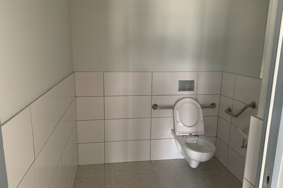 A clean, modern accessible bathroom featuring white wall tiles, a wall-mounted toilet, and stainless steel grab rails.