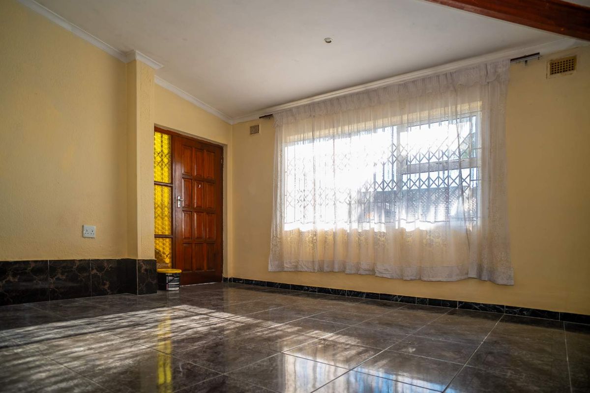 A bright living area featuring tiled flooring, a wooden entrance door, and a large window with security bars.