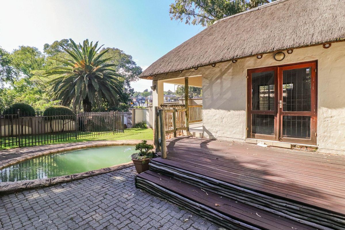 Rear exterior view featuring a thatched roof structure, timber deck, paved patio area, and a swimming pool with surrounding metal fencing.