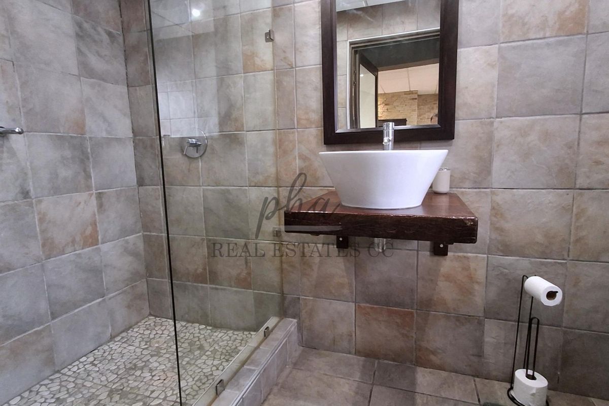 A modern bathroom featuring a glass-enclosed shower with pebble-style flooring, a vessel sink on a floating wooden vanity, and neutral wall tiling.