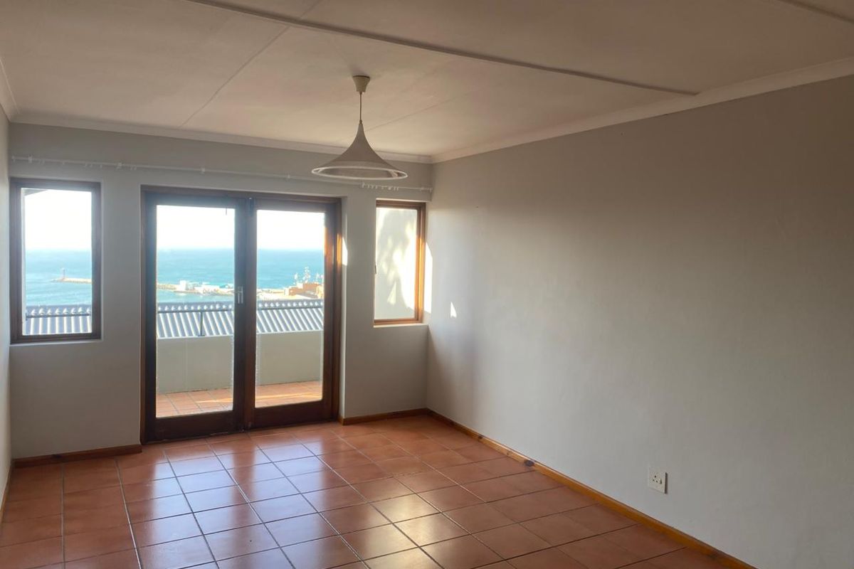 An empty living room featuring terracotta floor tiles, wooden framed glass doors leading to a balcony, and a scenic ocean view.