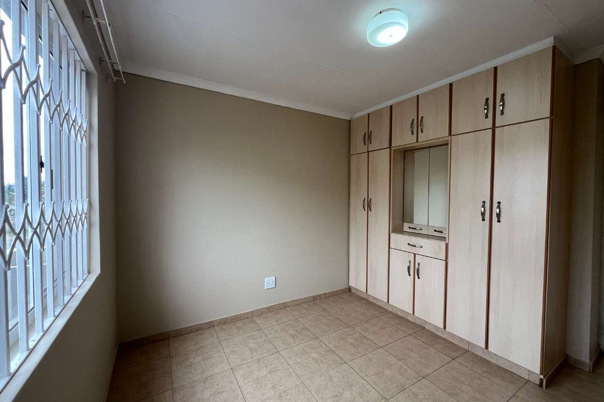 A clean, empty bedroom featuring neutral tiled flooring, built-in wooden cupboards with a vanity section, and a window fitted with a security gate.