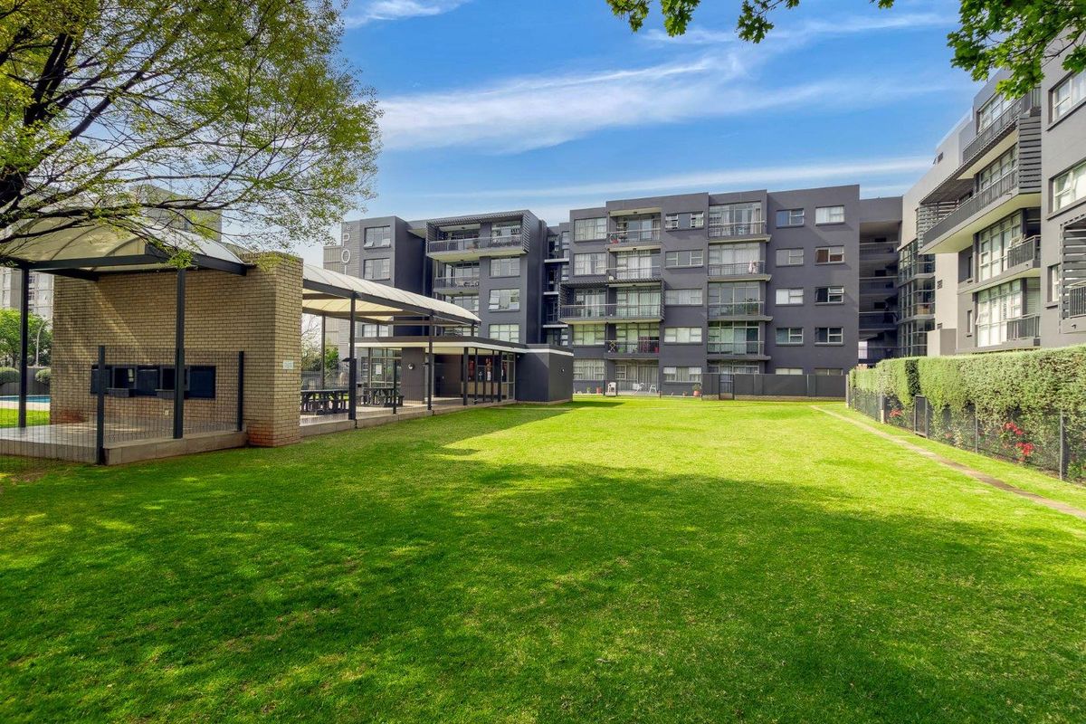 A well-maintained communal garden area featuring a covered braai facility and paved walkways, set against the backdrop of a modern apartment complex.