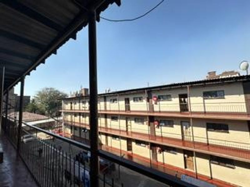 An open-air apartment balcony featuring metal railings and a corrugated roof, overlooking the adjacent residential building complex.