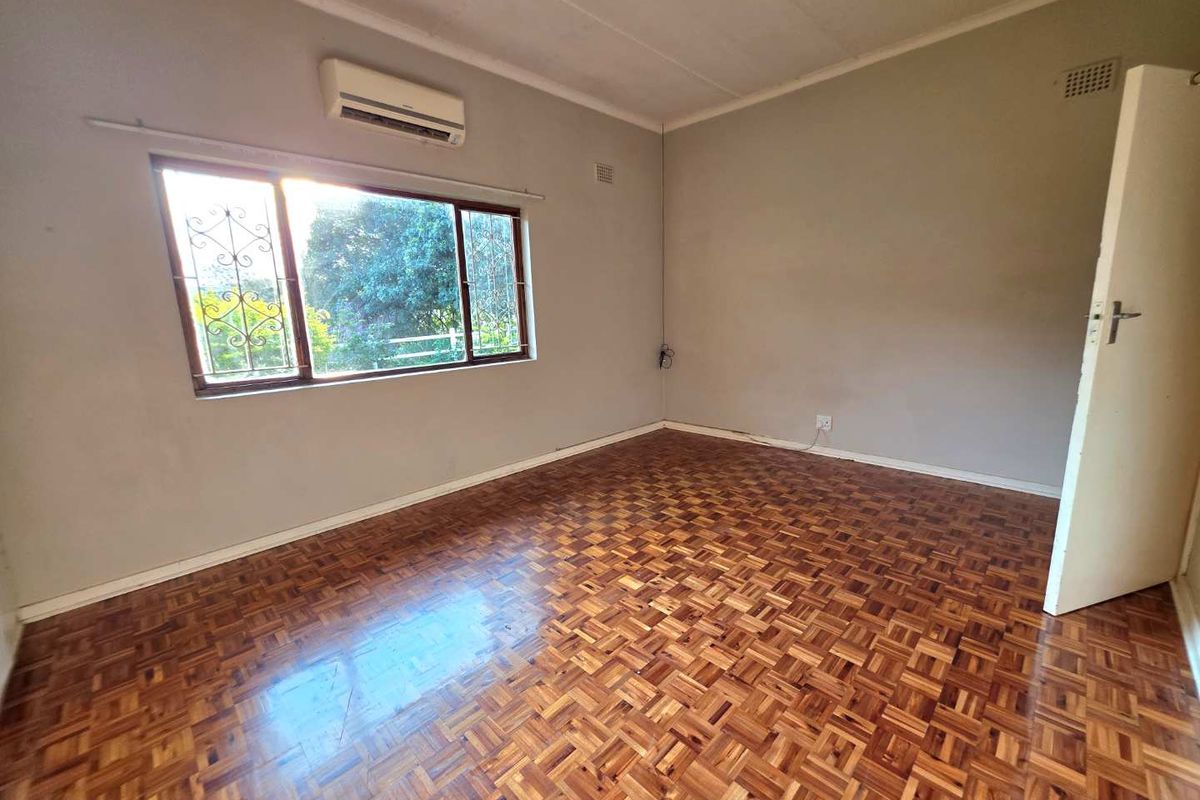 A bright, empty bedroom featuring traditional parquet wood flooring, a wall-mounted air conditioning unit, and large windows with security bars.