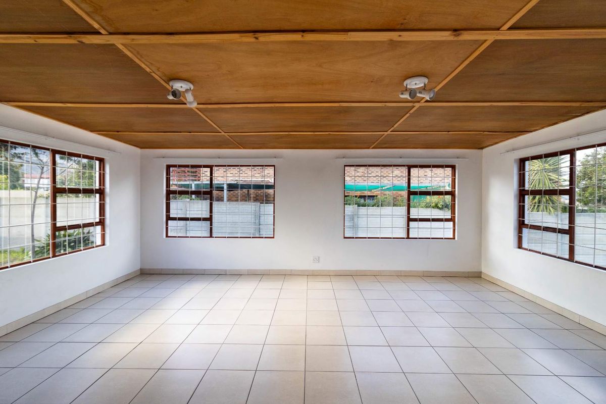 A spacious, empty living area featuring tiled flooring, a wooden panel ceiling, and large windows fitted with security bars.