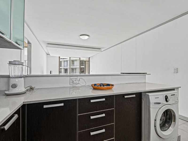 A functional kitchen area featuring dark cabinetry, stone countertops, and a dedicated space for a washing machine, opening into a bright living area.
