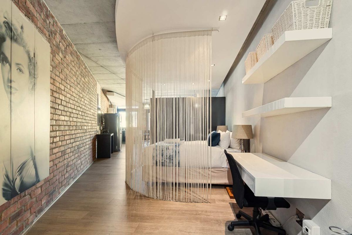Modern bedroom featuring an exposed brick wall, concrete ceiling, floating shelving, and a built-in desk, creating a stylish industrial aesthetic.