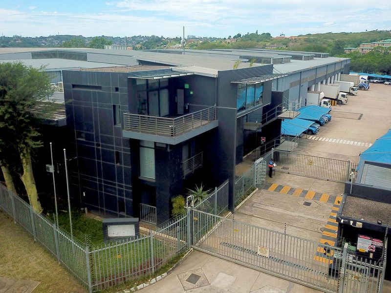 Aerial view of a modern industrial facility featuring a multi-level office block, secure gated entrance, paved parking area, and extensive warehouse space.
