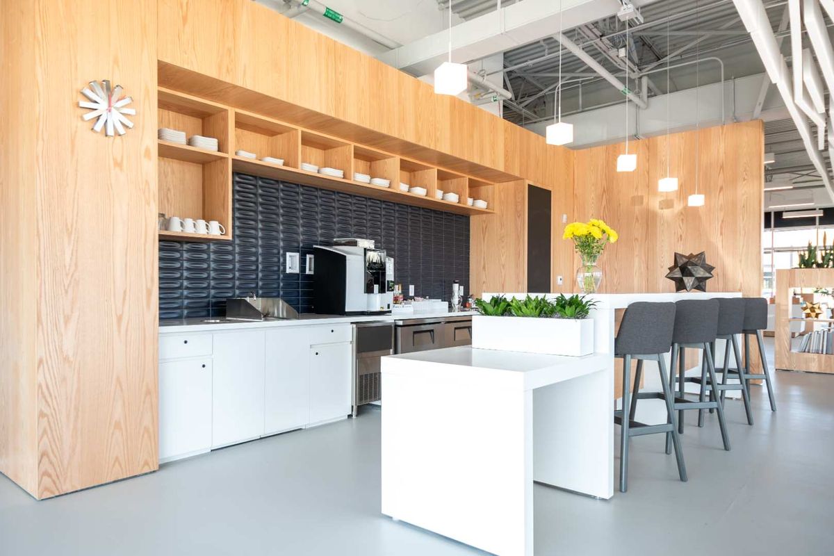Modern office kitchenette featuring light wood cabinetry, dark textured tile backsplash, integrated sink, and a clean white breakfast bar with seating.