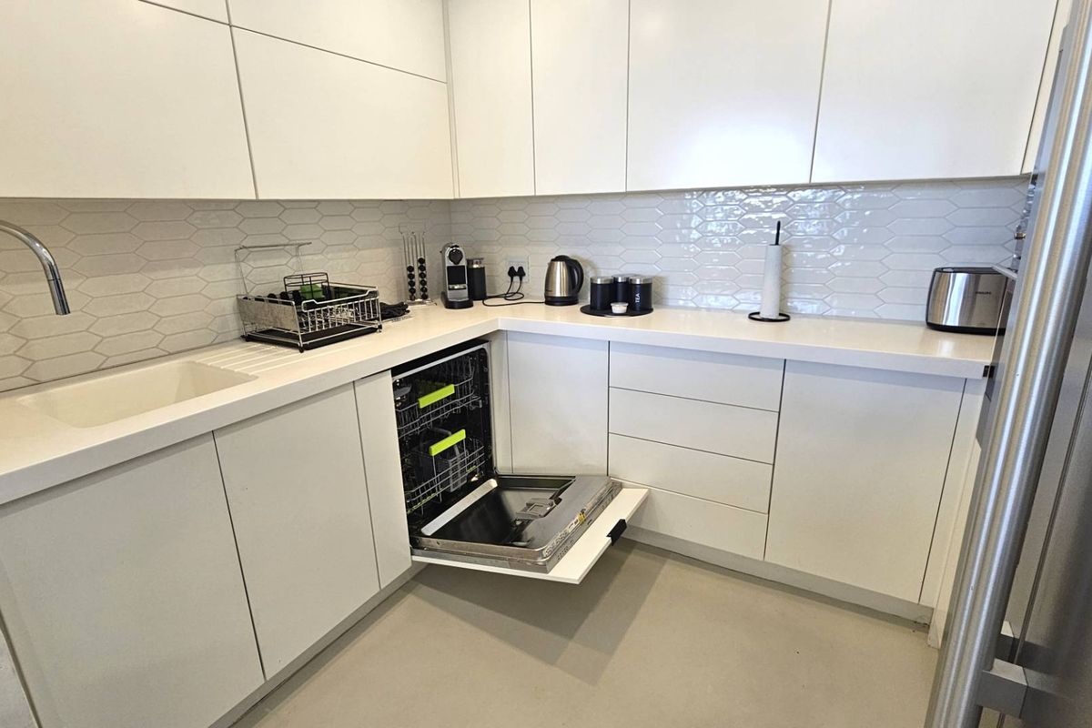 Modern kitchen featuring white handleless cabinetry, stone countertops, a tiled backsplash, and an integrated dishwasher.