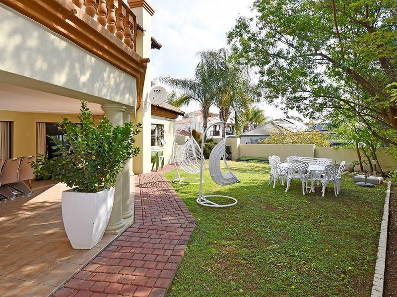 A rear garden view featuring a covered patio with tiled flooring, brick paved pathways, and a well-maintained lawn area.