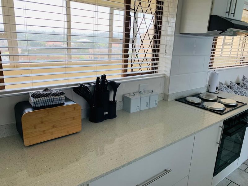 A modern kitchen featuring white cabinetry, quartz countertops, a built-in electric hob, oven, and extractor fan, complemented by a subway tile backsplash and window blinds.
