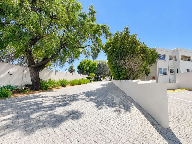 A well-maintained paved driveway leading to the apartment complex, featuring perimeter walls with electric fencing and established trees.