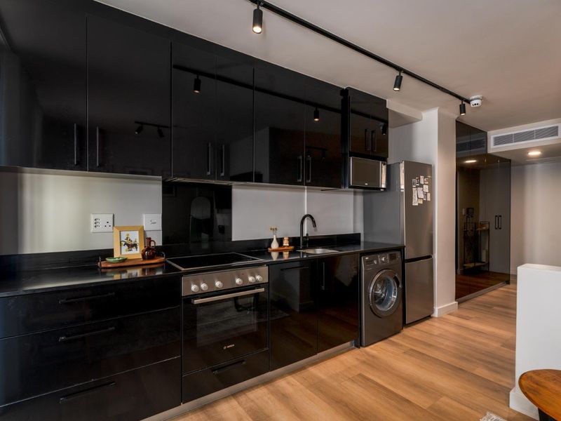 Modern kitchen featuring sleek black gloss cabinetry, integrated appliances, track lighting, and high-quality laminate wood flooring in a contemporary apartment setting.