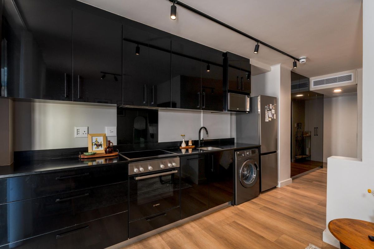 Modern kitchen featuring sleek black gloss cabinetry, integrated appliances, track lighting, and high-quality laminate wood flooring in a contemporary apartment setting.