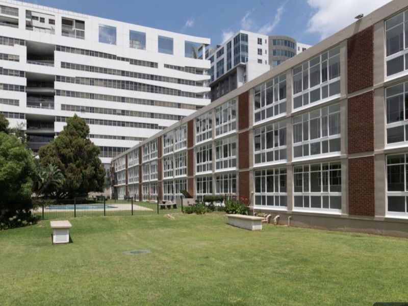 Exterior view of the apartment building featuring large windows, brick detailing, and access to a communal garden area with a swimming pool.