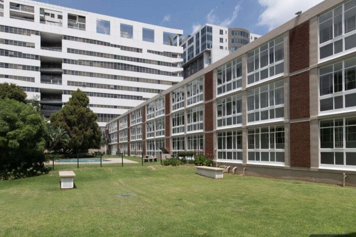Exterior view of the apartment building featuring large windows, brick detailing, and access to a communal garden area with a swimming pool.
